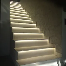 Nowoczesne schody z podświetleniem LED pod każdym stopniem, oświetlające ścieżkę w górę, ściana z dekoracyjną fakturą w tle.