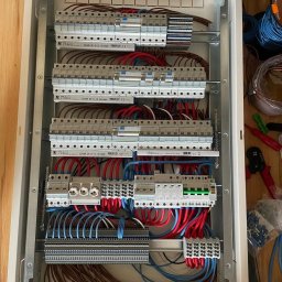 AWG SPÓŁKA Z OGRANICZONĄ ODPOWIEDZIALNOŚCIĄ - Rozdzielnica elektryczna w trakcie montażu, widoczne liczne przewody w kolorach niebieskim, czerwonym i brązowym, zabezpieczenia nadprądowe, listwy zaciskowe oraz narzędzia obok skrzynki.