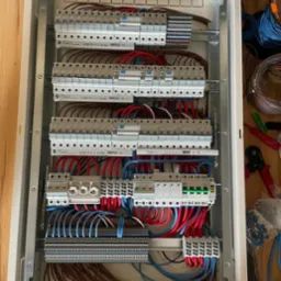 Rozdzielnica elektryczna w trakcie montażu, widoczne liczne przewody w kolorach niebieskim, czerwonym i brązowym, zabezpieczenia nadprądowe, listwy zaciskowe oraz narzędzia obok skrzynki.