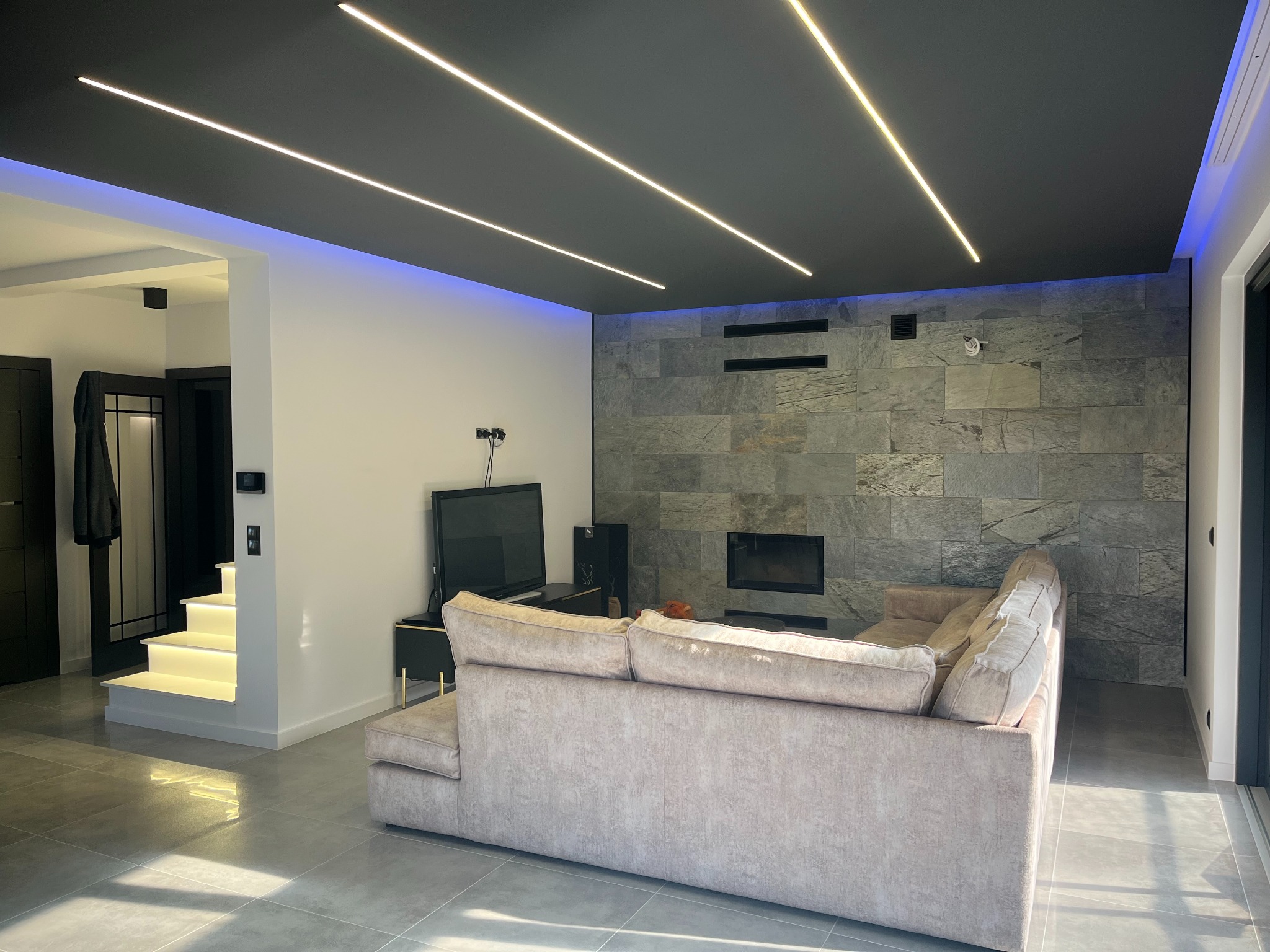 Nowoczesny salon z podświetlanym sufitem i schodami, linearnym oświetleniem LED na suficie, ściana z kamienia dekoracyjnego i podświetlane stopnie schodów.