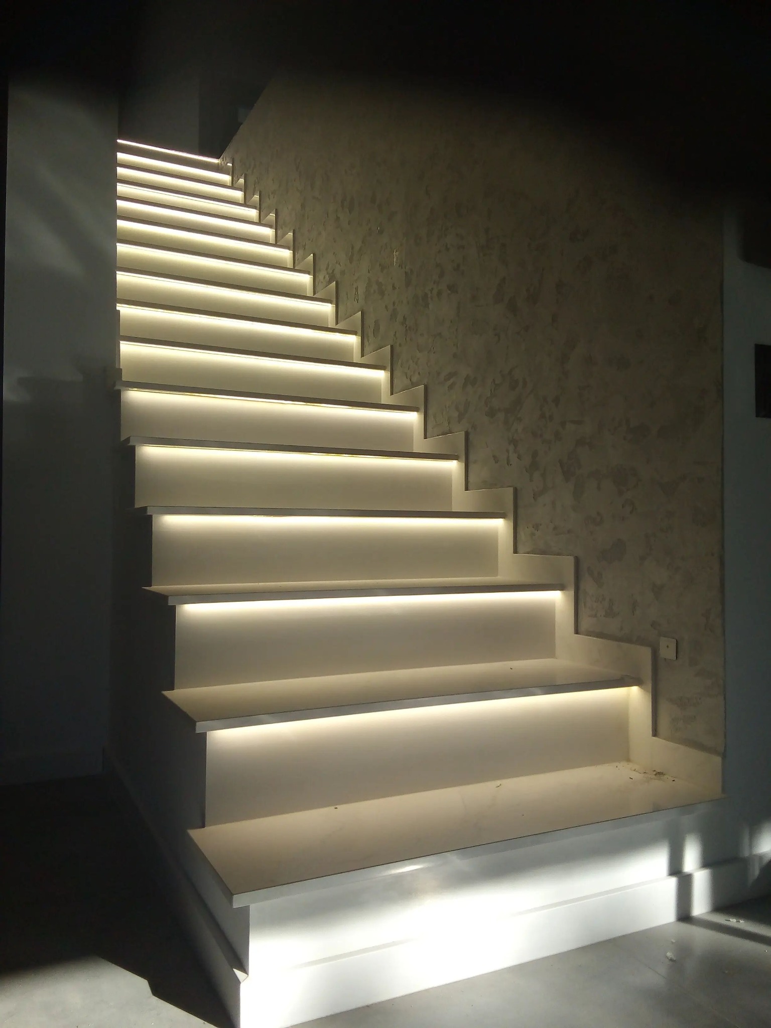 Nowoczesne schody z podświetleniem LED pod każdym stopniem, oświetlające ścieżkę w górę, ściana z dekoracyjną fakturą w tle.