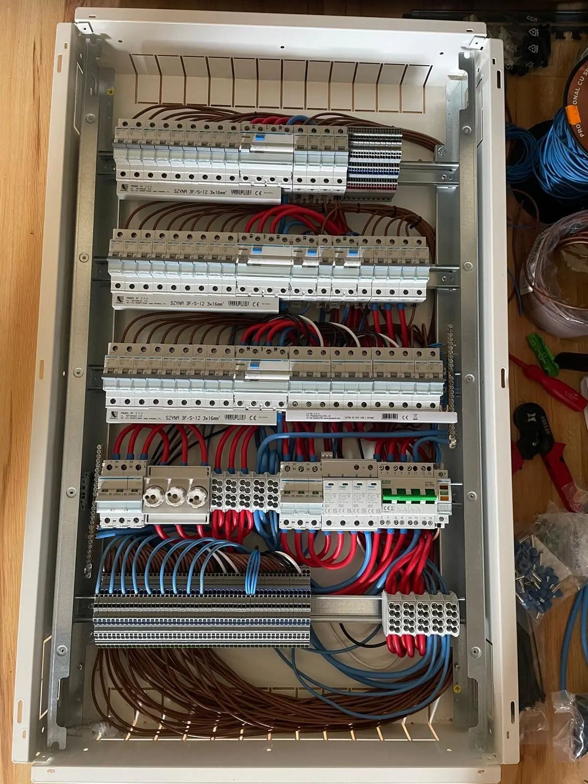 Rozdzielnica elektryczna w trakcie montażu, widoczne liczne przewody w kolorach niebieskim, czerwonym i brązowym, zabezpieczenia nadprądowe, listwy zaciskowe oraz narzędzia obok skrzynki.