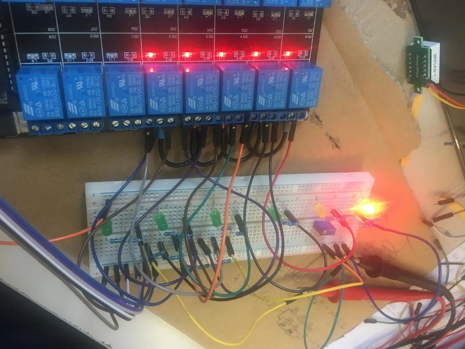 Testowanie obwodu elektrycznego z użyciem płytki stykowej, modułu przekaźników z diodami LED sygnalizującymi stan oraz multimetru z czerwoną sondą, widoczne liczne przewody połączeniowe.
