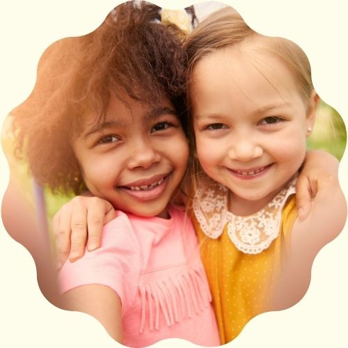 Uśmiechnięte dziewczynki, jedna w różowej bluzce z frędzlami, druga w żółtej sukience z koronkowym kołnierzykiem, obejmują się na tle zieleni.