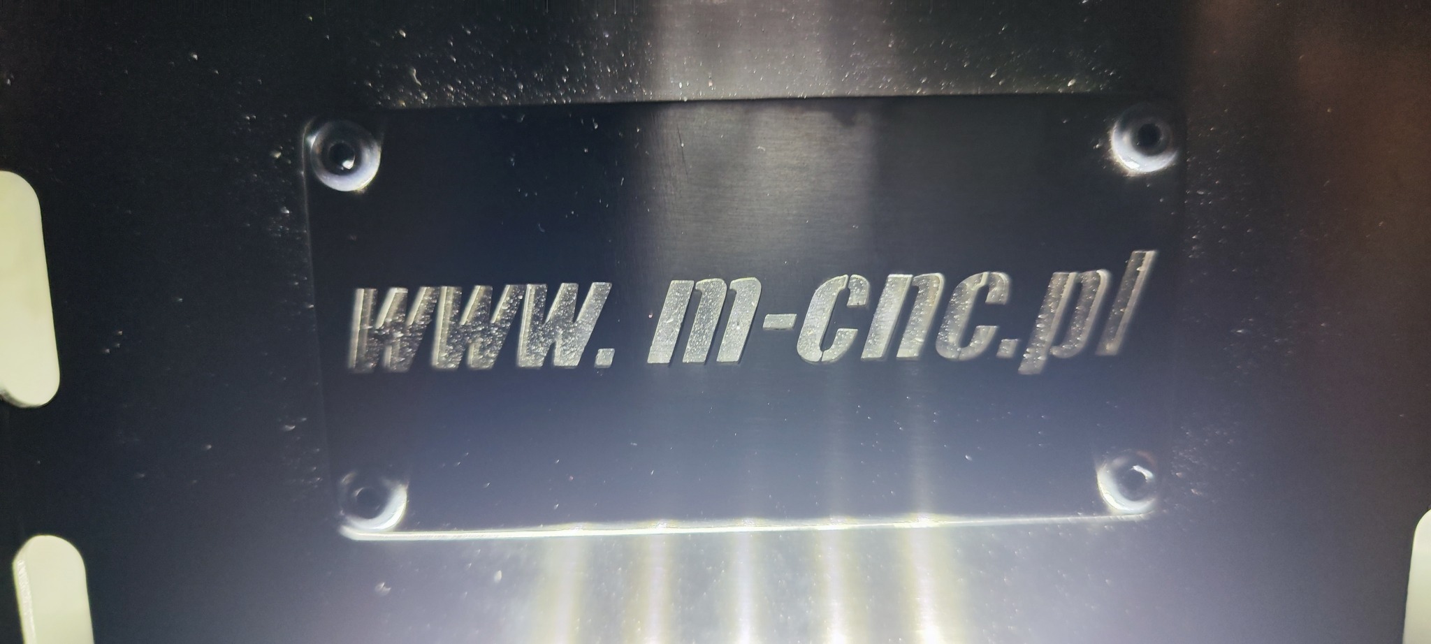 Czarna metalowa tabliczka z wygrawerowanym adresem strony internetowej www.m-cnc.pl, przymocowana czterema śrubami.
