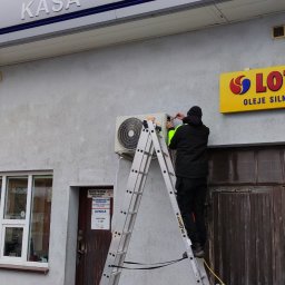 BARNET Seweryn Tyborski - Montaż klimatyzatora zewnętrznego na ścianie budynku stacji benzynowej Lotos przez technika na drabinie.