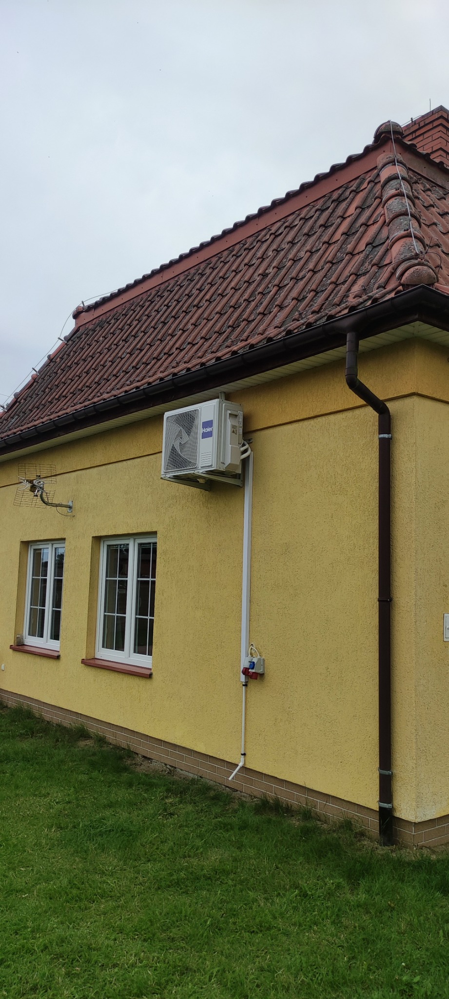 Zewnętrzna jednostka klimatyzacji Haier zamontowana na żółtej elewacji domu z brązowym dachem z dachówki ceramicznej, widoczna instalacja elektryczna i rura odprowadzająca skropliny.
