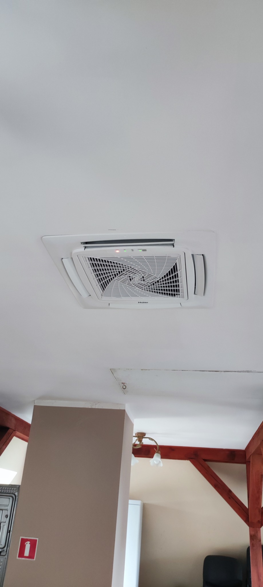 Kasetonowa jednostka klimatyzacyjna Haier zamontowana w suficie, widoczne diody sygnalizacyjne i kratka wentylacyjna, w tle fragment pomieszczenia z drewnianymi elementami konstrukcyjnymi.