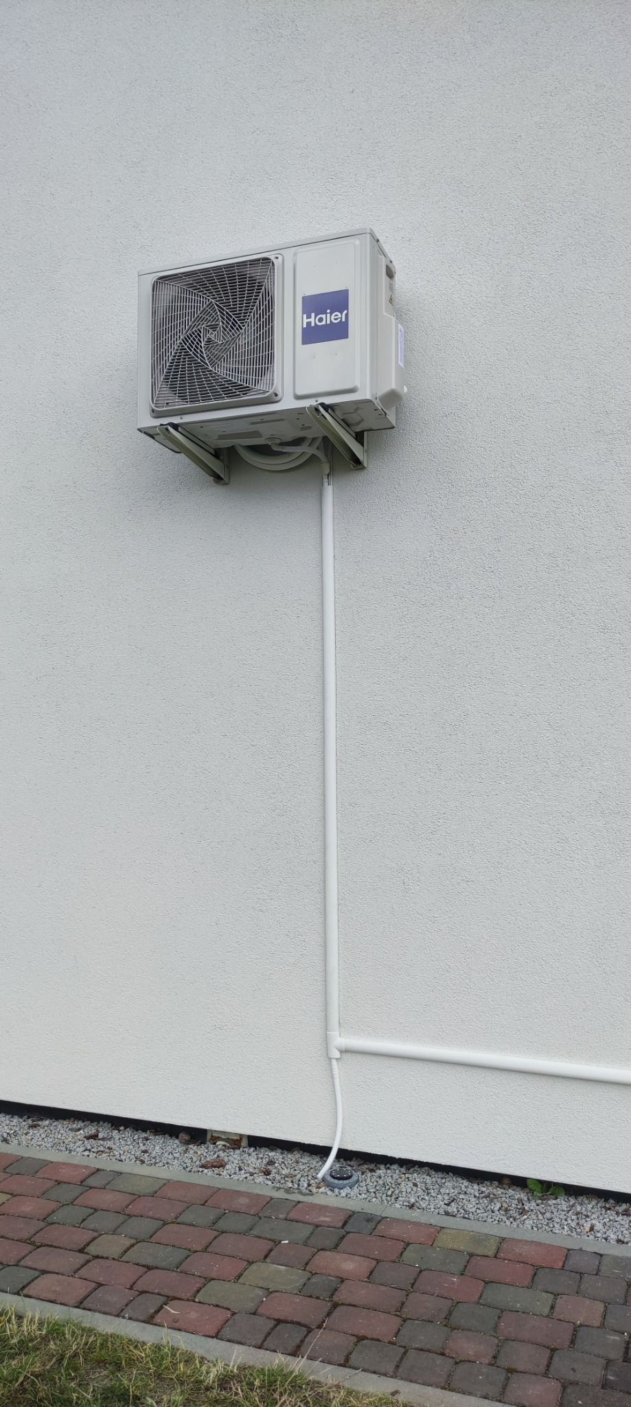 Biała jednostka zewnętrzna klimatyzatora Haier zamontowana na jasnej, teksturowanej ścianie budynku, z widocznymi rurami odprowadzającymi skropliny i przewodami, prowadzącymi wzdłuż ściany...