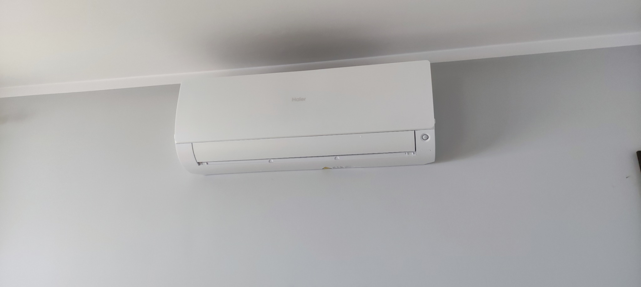 Zamontowana na ścianie, biała jednostka wewnętrzna klimatyzacji Haier w pomieszczeniu o szarych ścianach.