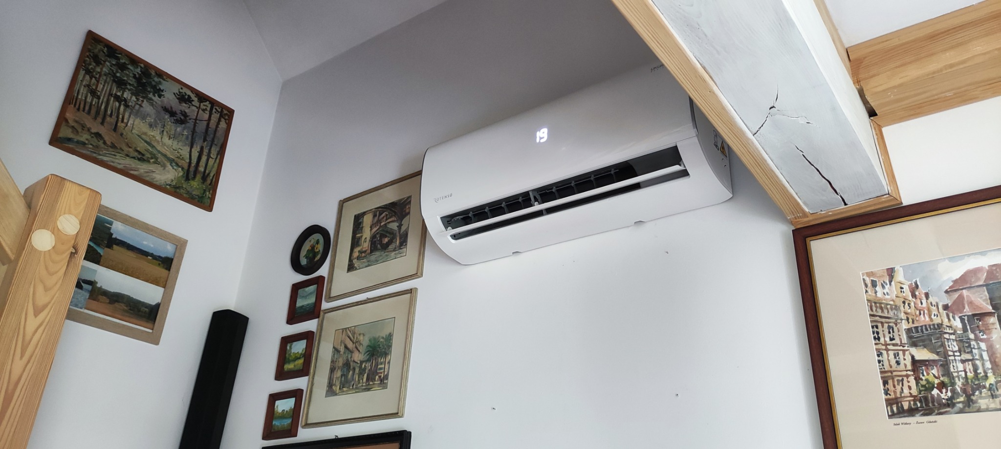 Biały klimatyzator ścienny marki Cotenso, wyświetlający temperaturę 19 stopni Celsjusza, zamontowany w pomieszczeniu z drewnianą belką sufitową i galerią obrazów w ozdobnych ramach.