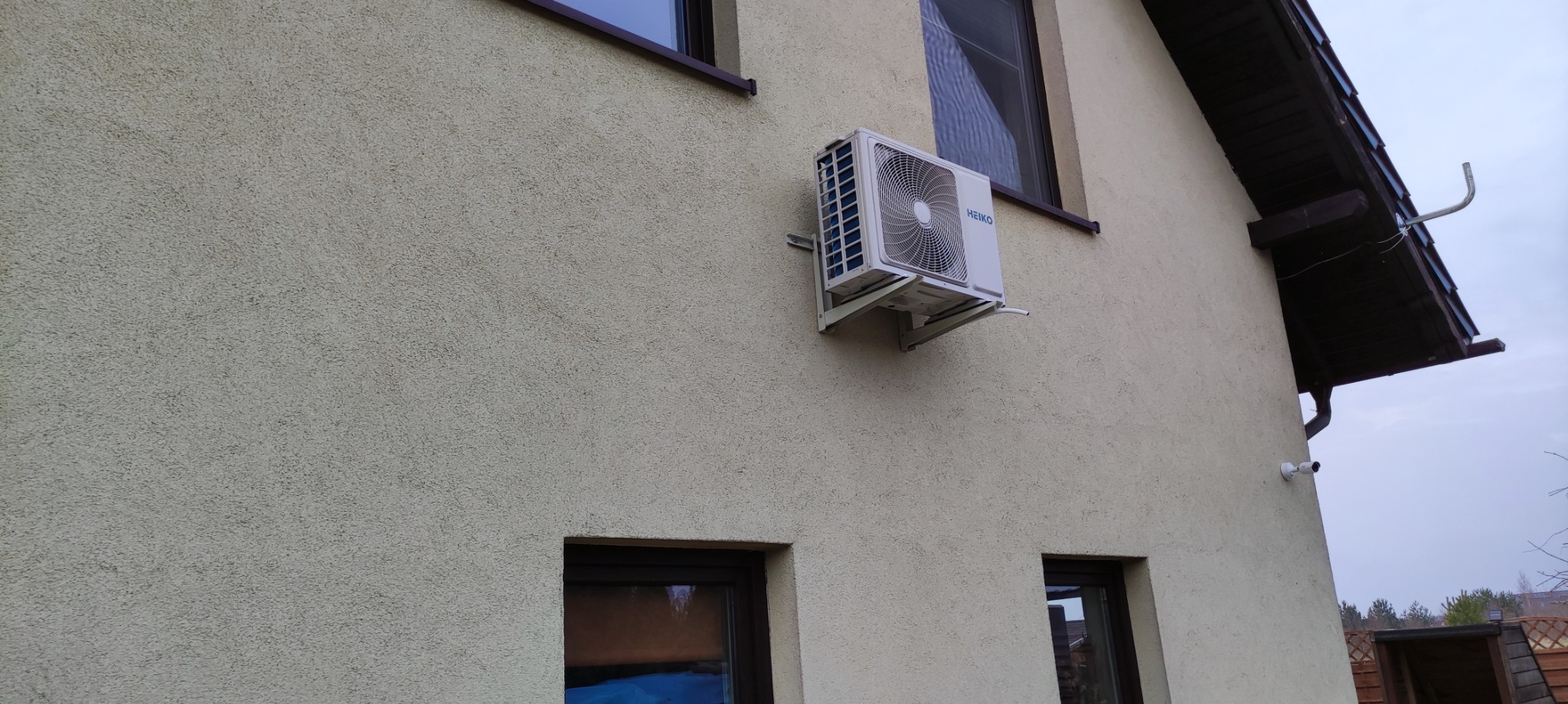 Biała jednostka zewnętrzna klimatyzatora marki Heiko zamontowana na kremowej elewacji domu, widoczna instalacja odprowadzająca skropliny, okna z brązowymi ramami i fragment dachu.