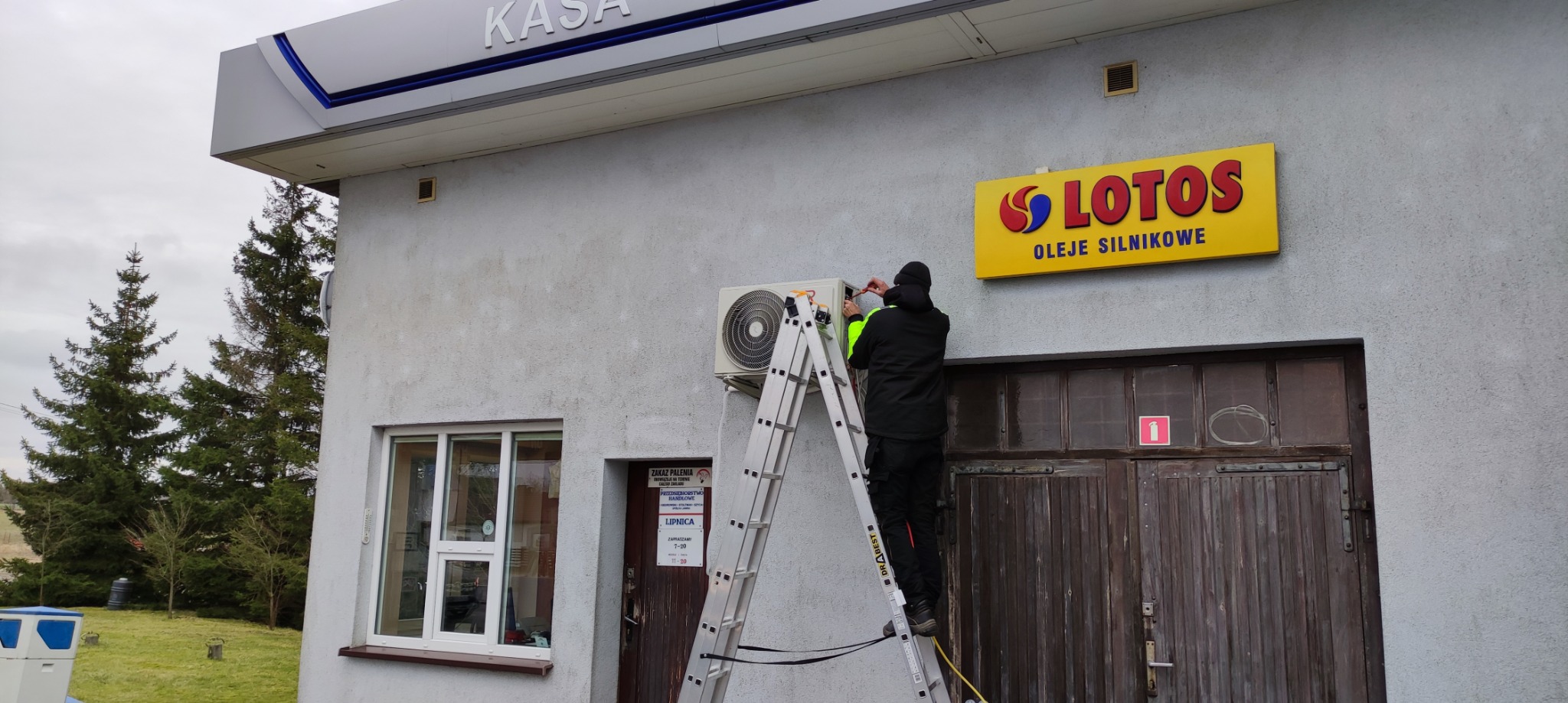 Montaż klimatyzatora zewnętrznego na ścianie budynku stacji benzynowej Lotos przez technika na drabinie.