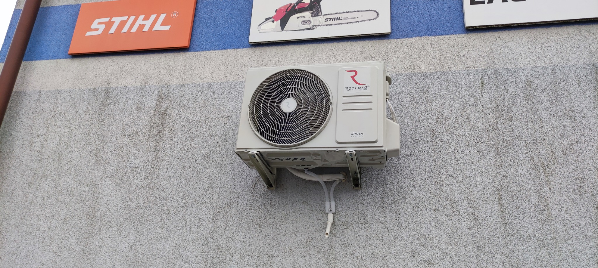 Zamontowany na zewnątrz budynków klimatyzator marki Rotenso, widoczne rury odprowadzające skropliny, nad urządzeniem wiszą tablice reklamowe Stihl.