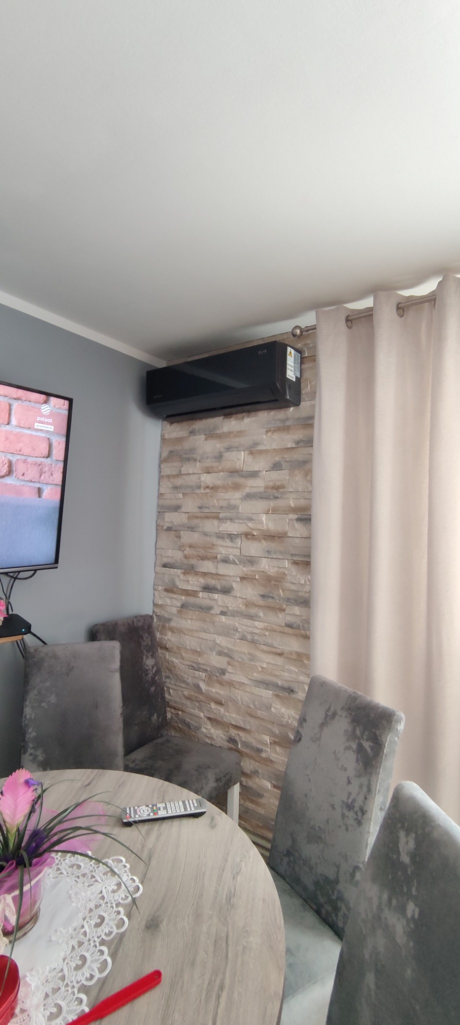 Czarny klimatyzator ścienny zamontowany nad dekoracyjną ścianą z kamienia w nowoczesnym salonie z szarymi krzesłami i okrągłym stołem.