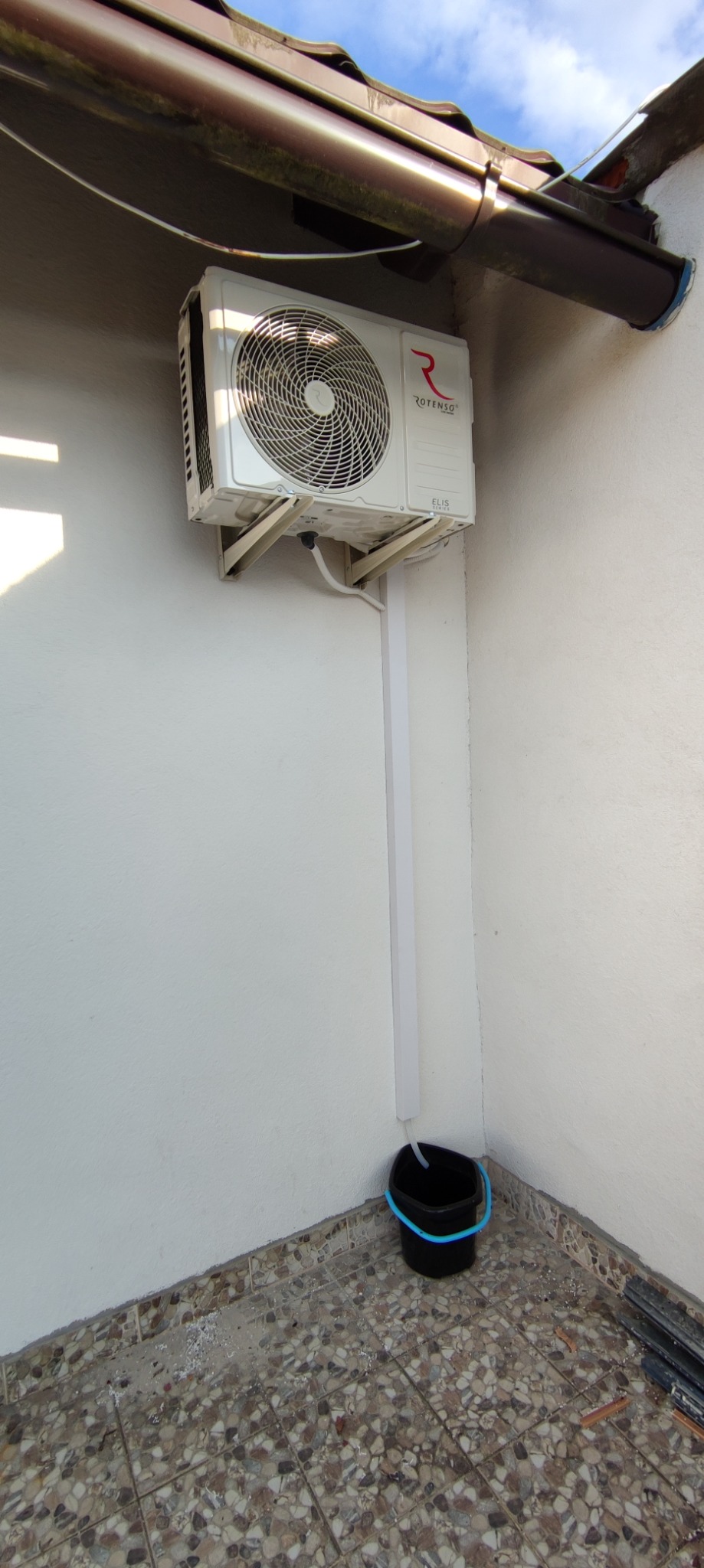 Biała jednostka zewnętrzna klimatyzatora marki Rotenso zamontowana na ścianie budynku, z odpływem wody skierowanym do czarnego wiadra z niebieskim uchwytem, widoczne ślady prac montażowych...