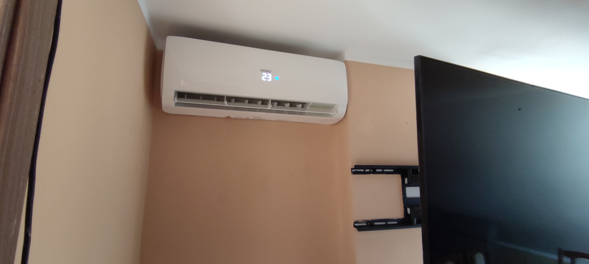 Biały klimatyzator Heiko z wyświetlaną temperaturą 23 stopnie Celsjusza, zamontowany na beżowej ścianie obok uchwytu do telewizora i częściowo widocznego czarnego ekranu telewizora.