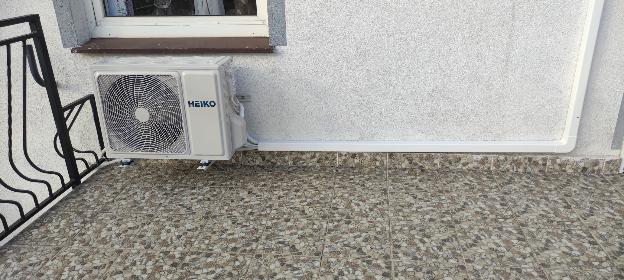Biały klimatyzator marki HEIKO zamontowany na zewnątrz budynku, z widocznymi elementami instalacji i brązowym parapetem okna w tle.