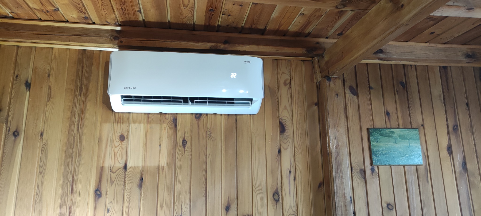 Biały klimatyzator marki Rotenso zamontowany na drewnianej ścianie w domu z boazerią, wyświetlający temperaturę 22 stopnie.