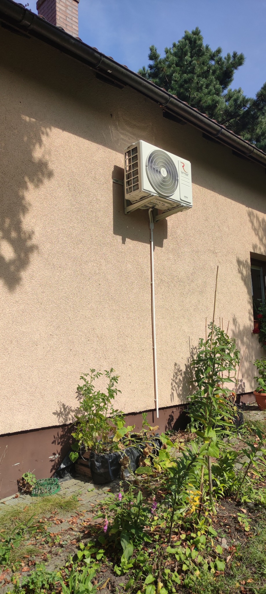 Biała jednostka zewnętrzna klimatyzacji zamontowana na elewacji domu z beżowym tynkiem, widoczne rury odprowadzające skropliny, w tle ogródek z roślinami.