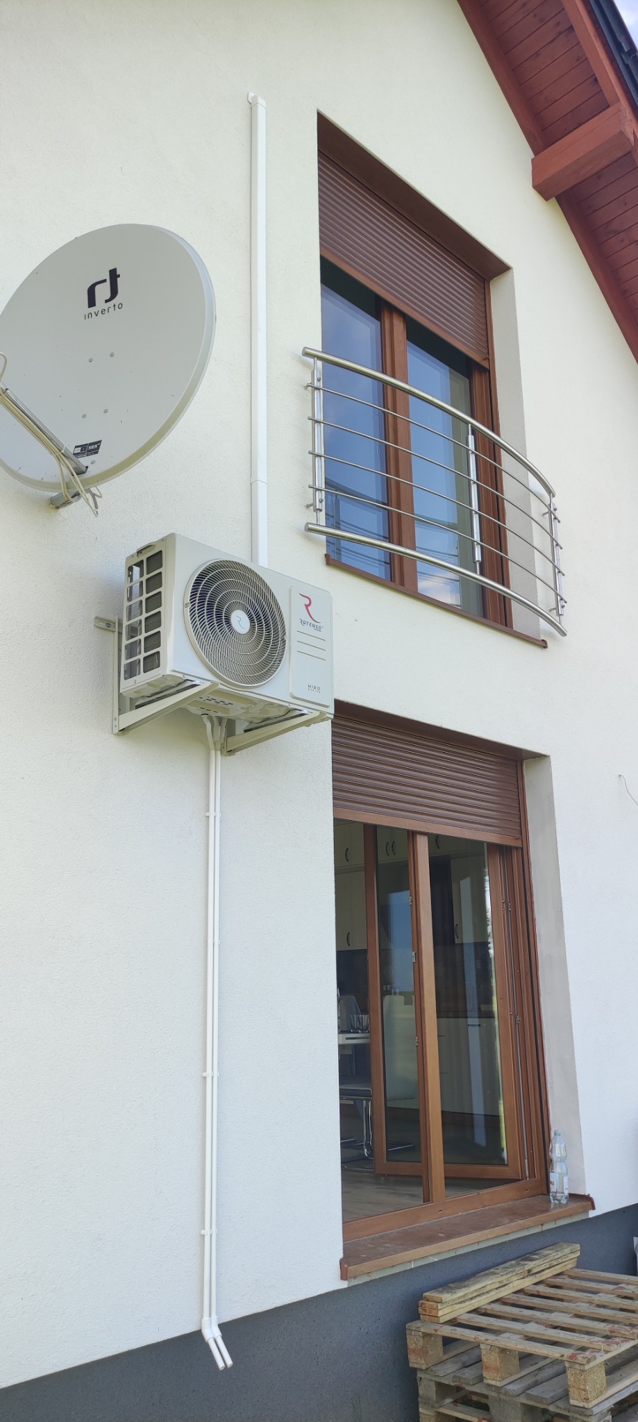 Zewnętrzna jednostka klimatyzacji Rotenso Hiro zamontowana na białej ścianie domu, widoczne rury odprowadzające skropliny, obok antena satelitarna i brązowe rolety okienne.
