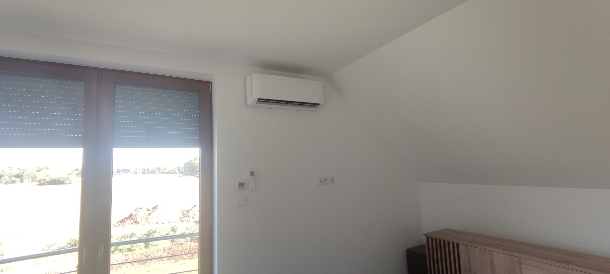 Biała jednostka klimatyzacyjna zamontowana na ścianie w jasnym pokoju, widoczny fragment okna z roletami i mebel w tle.