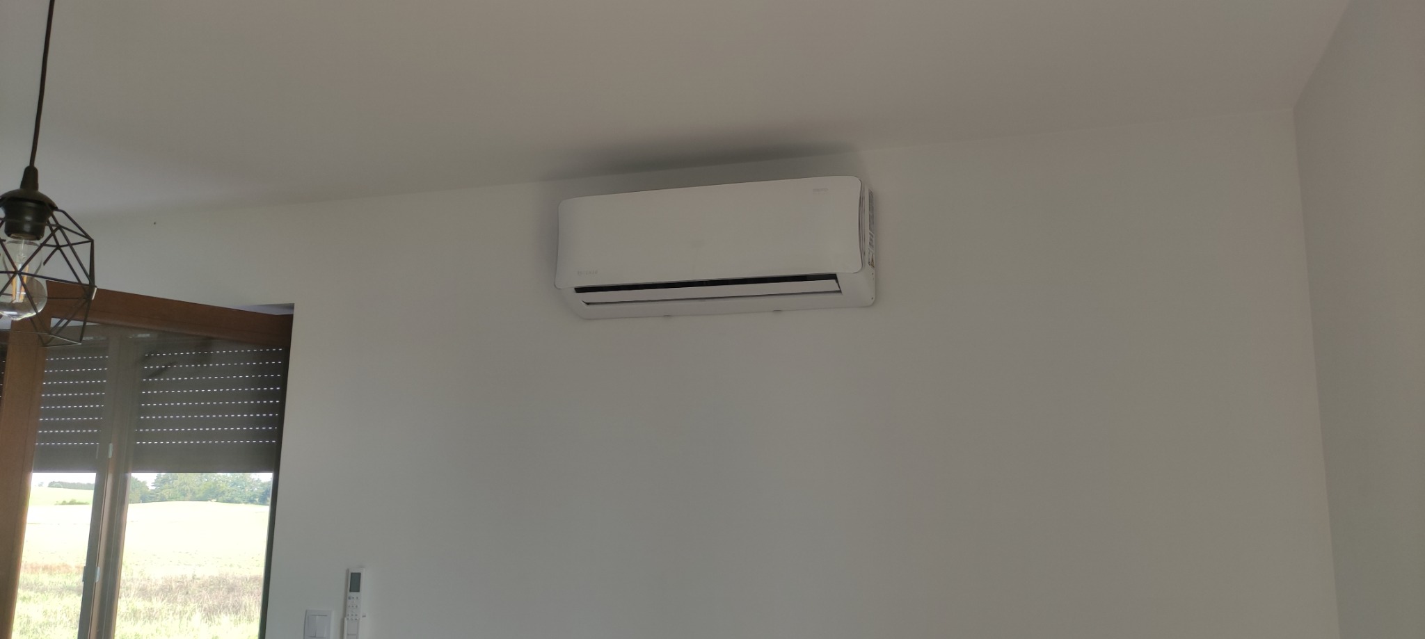 Biały klimatyzator ścienny w nowoczesnym wnętrzu, widoczny fragment okna z roletami zewnętrznymi i lampa wisząca.