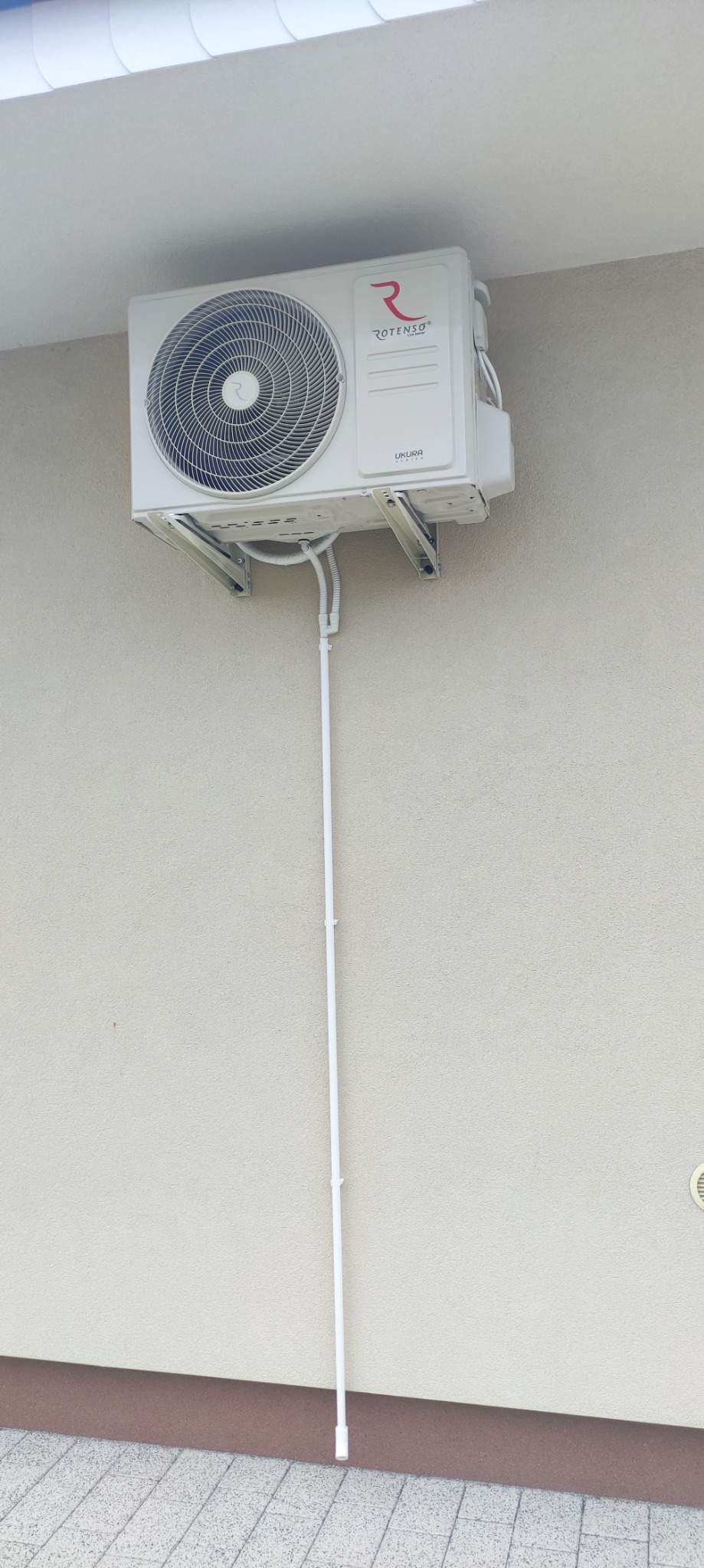 Biała jednostka zewnętrzna klimatyzacji Rotenso UKURA zamontowana na ścianie budynku z widocznymi rurami odprowadzającymi skropliny.