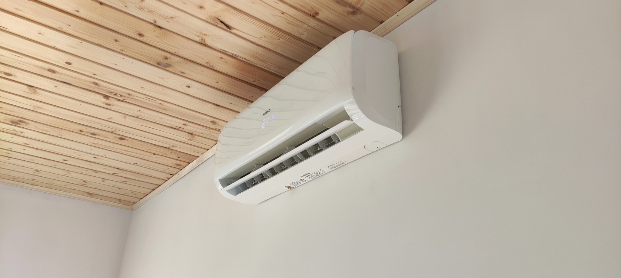 Biały klimatyzator marki HEIKO zamontowany na ścianie w pomieszczeniu z drewnianym panelem sufitowym.