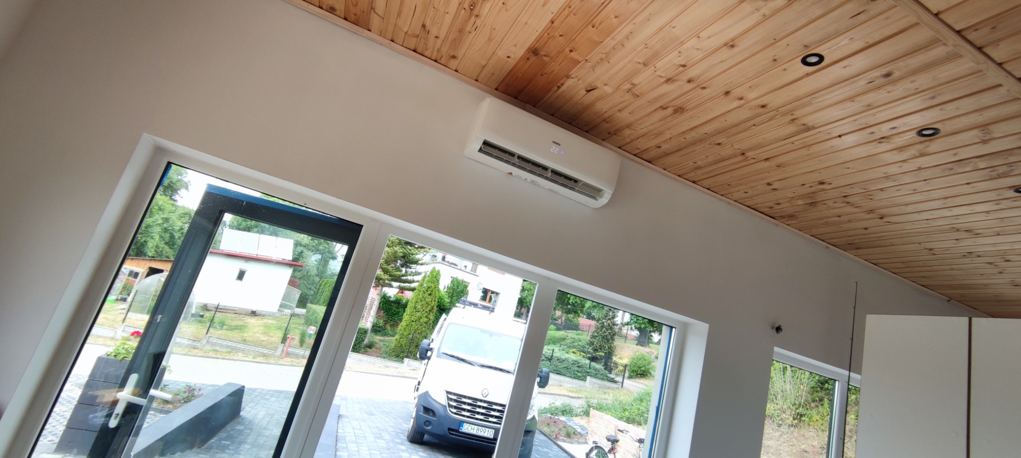 Biały klimatyzator zamontowany na ścianie w pomieszczeniu z drewnianym sufitem, widok przez okno na zaparkowany samochód dostawczy.