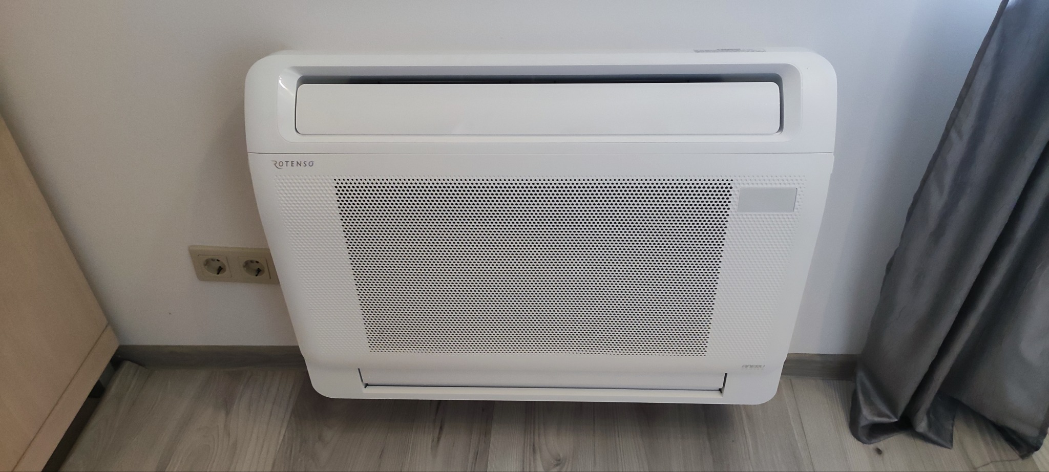 Biały klimatyzator przypodłogowy marki Rotenso zamontowany przy ścianie z widocznym gniazdkiem elektrycznym i fragmentem zasłony.
