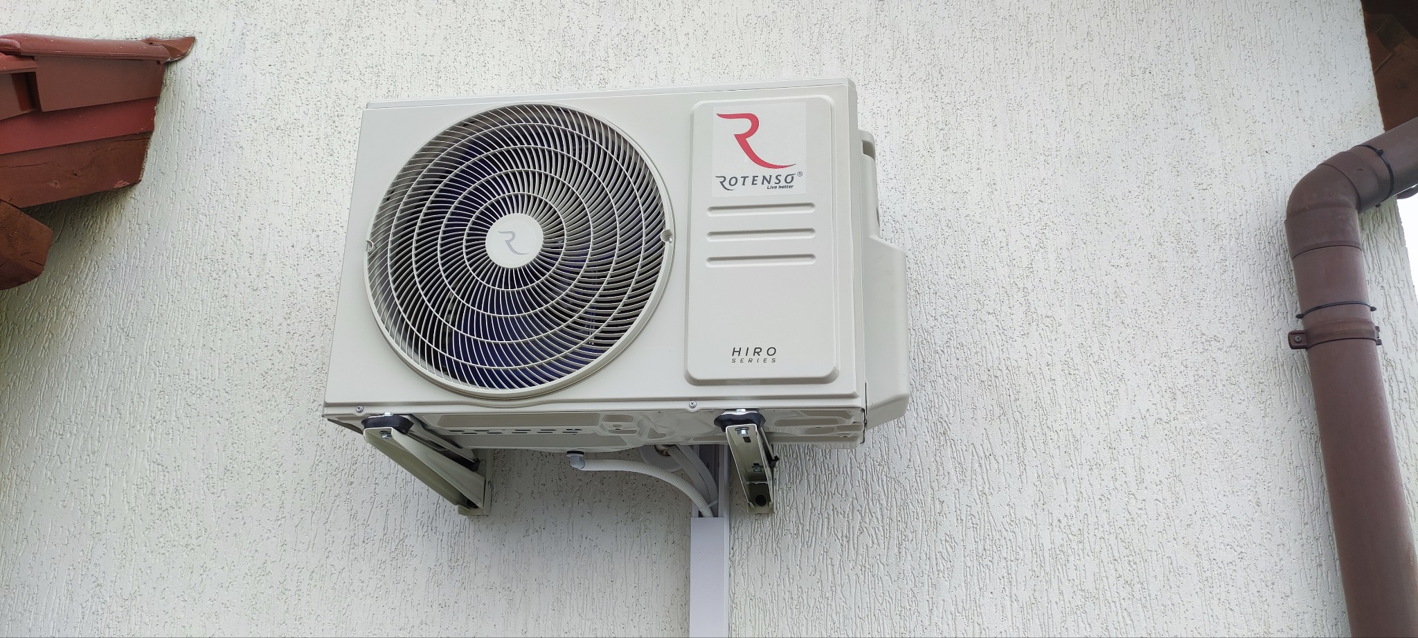 Biała jednostka zewnętrzna klimatyzatora Rotenso Hiro Series zamontowana na kremowej elewacji budynku, widoczna rynna w kolorze brązowym.