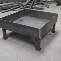 Surowa, spawana konstrukcja metalowa z kwadratową misą na czterech nogach, w tle nieporządek metalowych profili.