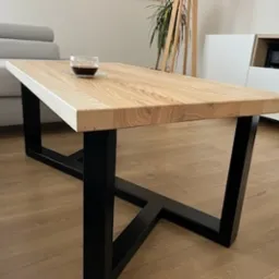 Stolik kawowy z blatem z jasnego drewna i czarnymi, metalowymi nogami w kształcie litery V, na blacie szklanka z napojem, w tle sofa i biała komoda.