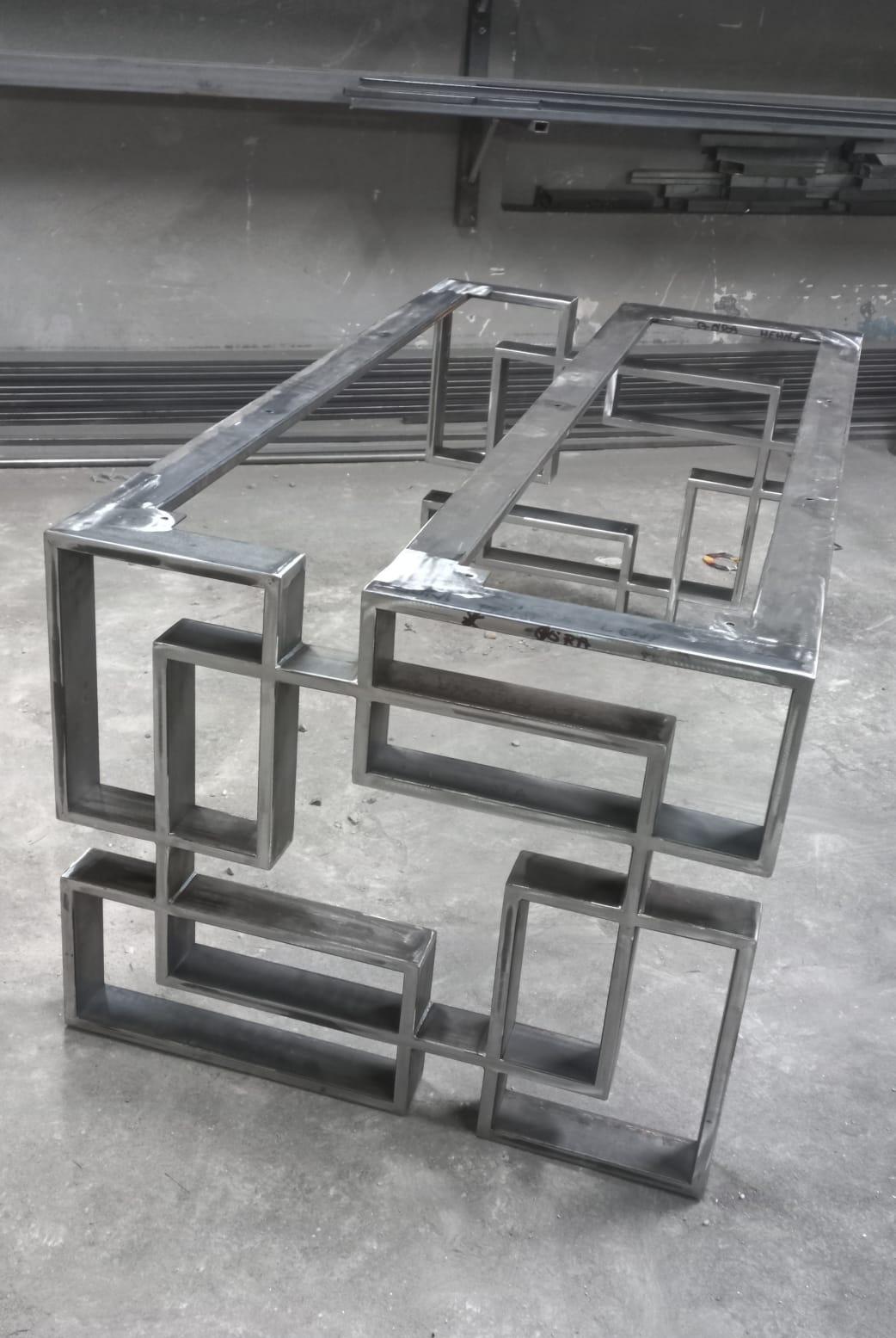 Metalowa konstrukcja stolika kawowego z profili kwadratowych, surowe spawy, widoczne niedoskonałości, szlifowane punkty łączeń, w tle profile stalowe.