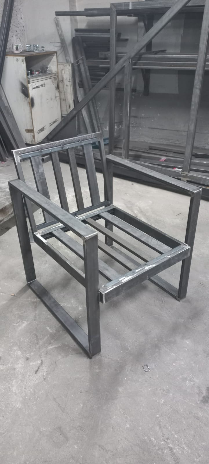 Metalowy szkielet krzesła z profili kwadratowych, spawane łączenia, surowy wygląd, warsztat w tle.