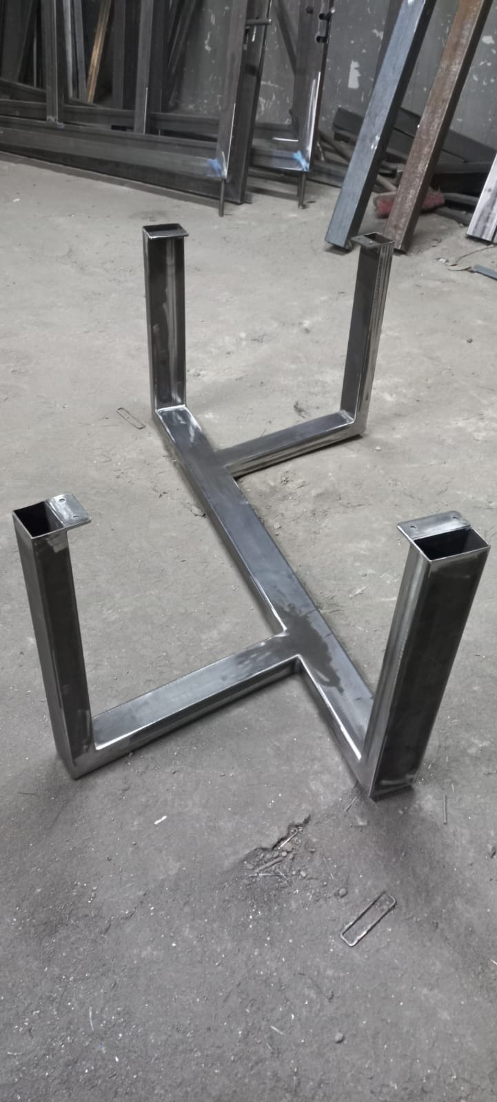 Metalowa konstrukcja ramy stołu z profili kwadratowych, spawana, surowa, leżąca na betonowej posadzce warsztatu, widoczne ślady spawania.