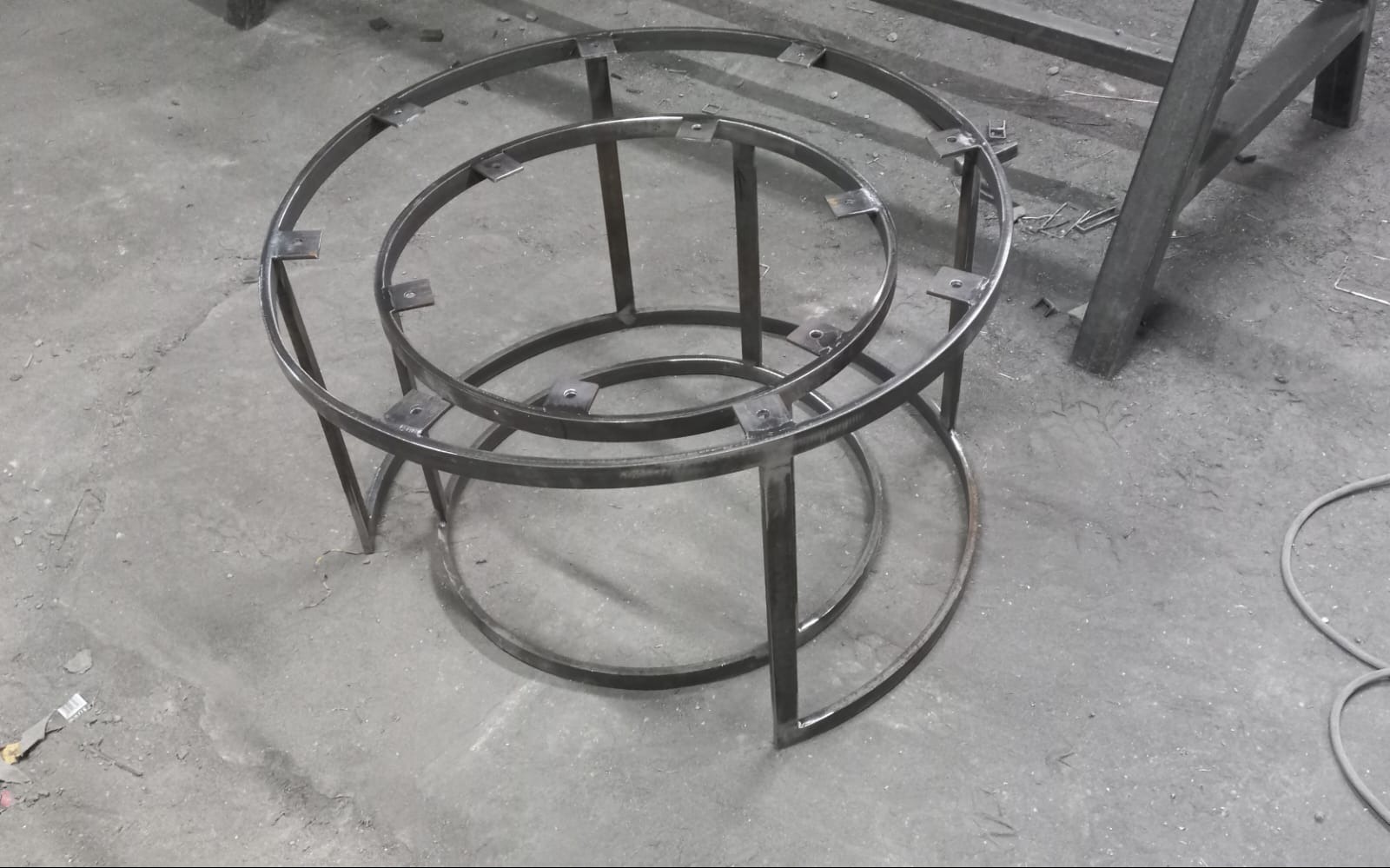 Metalowa konstrukcja składająca się z dwóch okręgów połączonych pionowymi wspornikami i małymi kwadratowymi płytkami, prawdopodobnie element większej konstrukcji, widoczne ślady obróbki metalu.