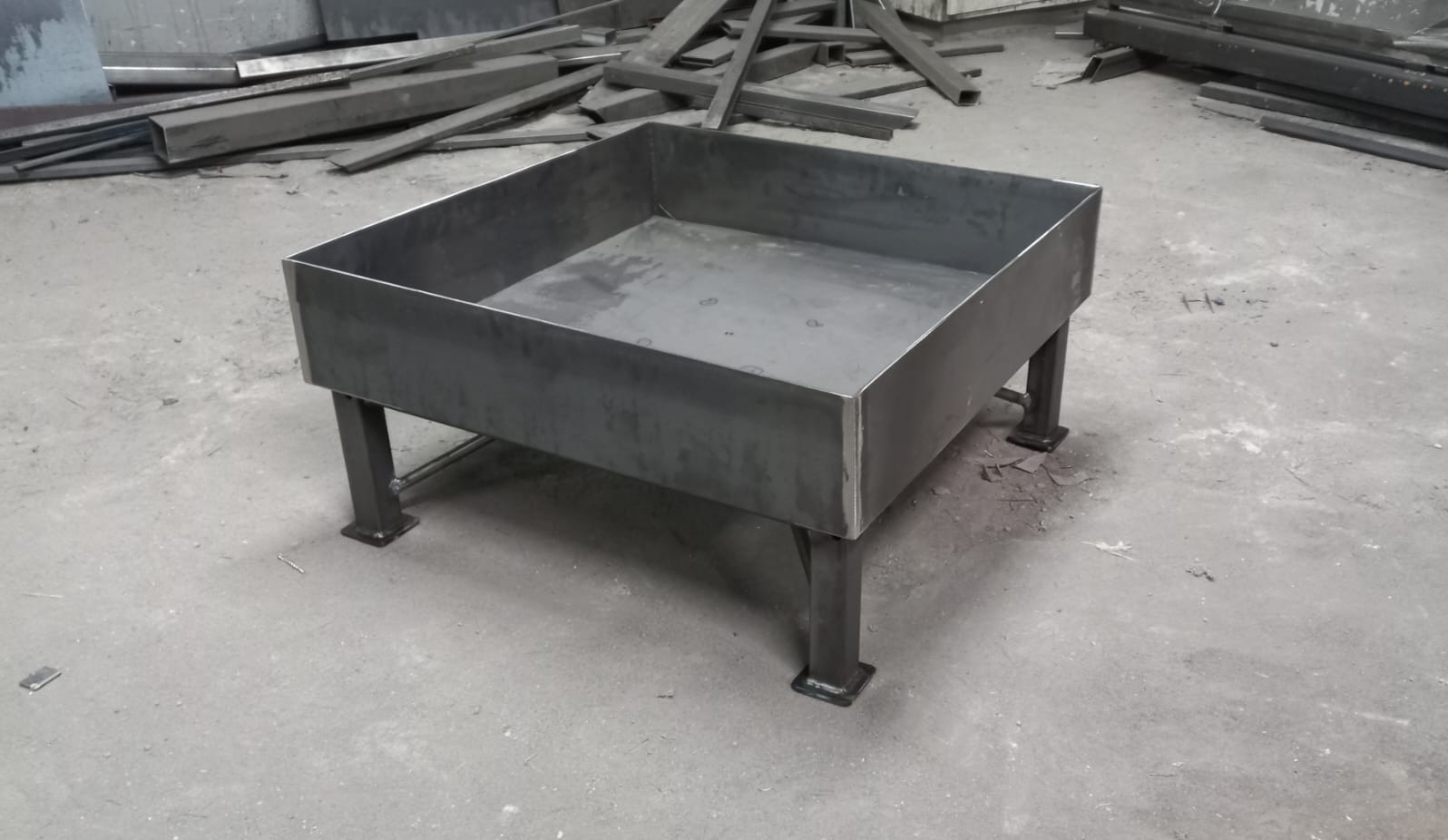Surowa, spawana konstrukcja metalowa z kwadratową misą na czterech nogach, w tle nieporządek metalowych profili.