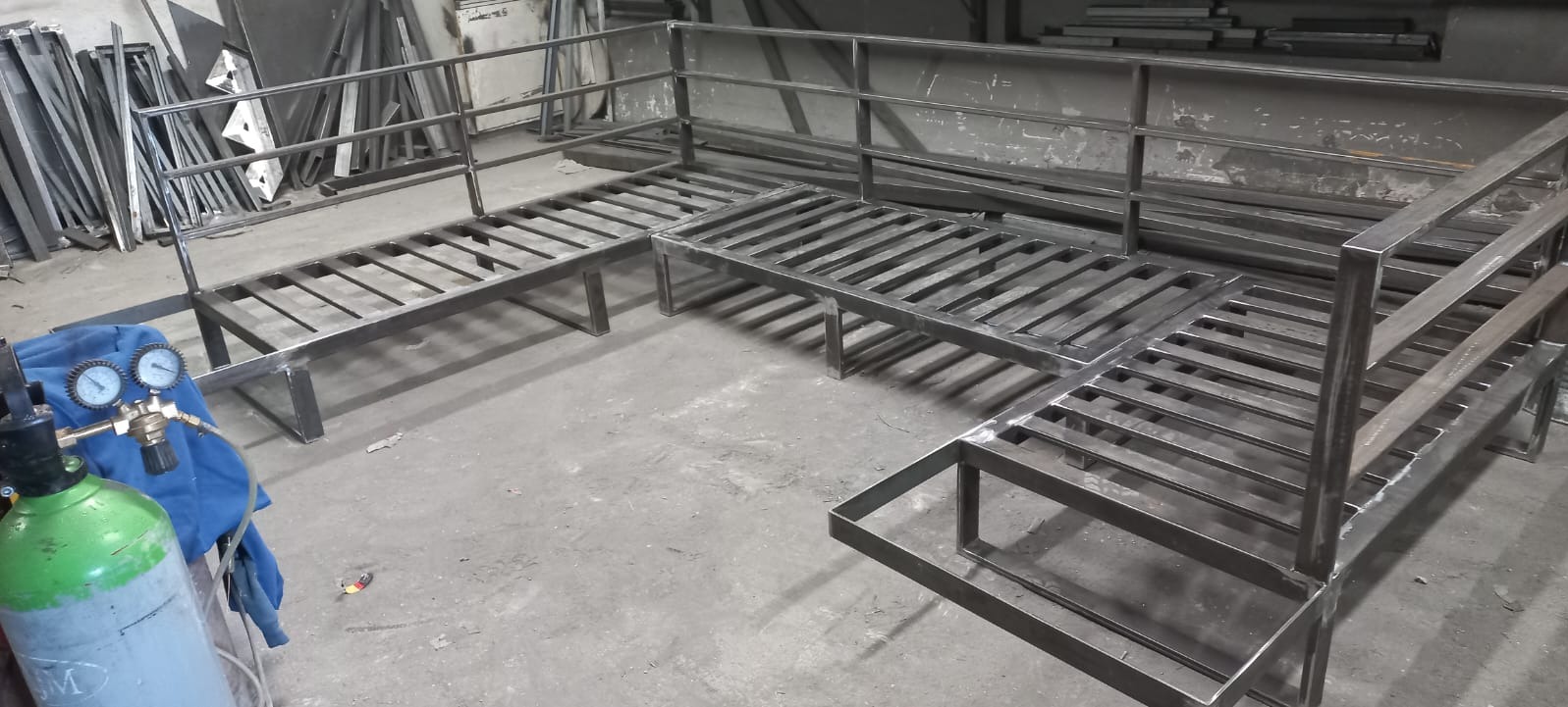 Konstrukcja metalowa w trakcie spawania, przypominająca ramę sofy ogrodowej, z widocznym butlą spawalniczą w tle, surowy beton na podłodze.