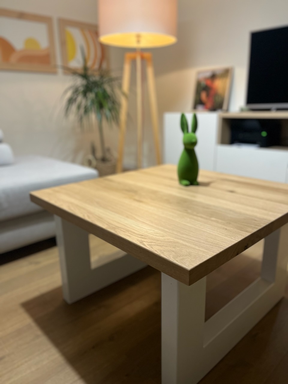 Kwadratowy stolik kawowy z jasnego drewna z białymi nogami w nowoczesnym salonie, na blacie zielona figurka zająca.