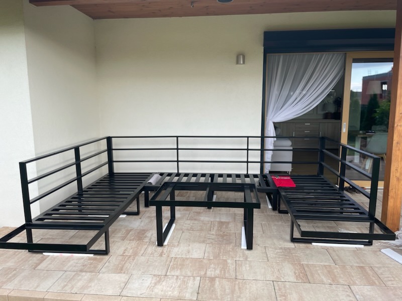 Metalowa konstrukcja narożnej sofy i stolika kawowego, czarne profile, białe podkładki, częściowo zmontowana, taras z jasnych płytek, widok na okno tarasowe z zasłonami.