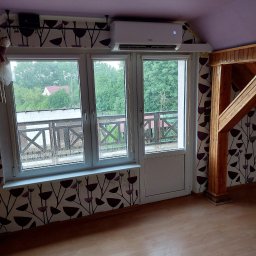 ELEKTROSERWIS Paweł Waligórski - Biała jednostka klimatyzacyjna zamontowana nad oknem balkonowym w pokoju z wzorzystą tapetą i drewnianą konstrukcją. Widok z okna na zewnątrz.