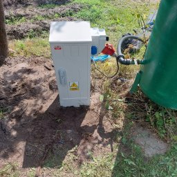 ELEKTROSERWIS Paweł Waligórski - Zewnętrzna szafa elektryczna z ostrzeżeniem o wysokim napięciu, obok zielonego zbiornika, na trawiastym terenie z widocznymi przewodami i elementami instalacji.