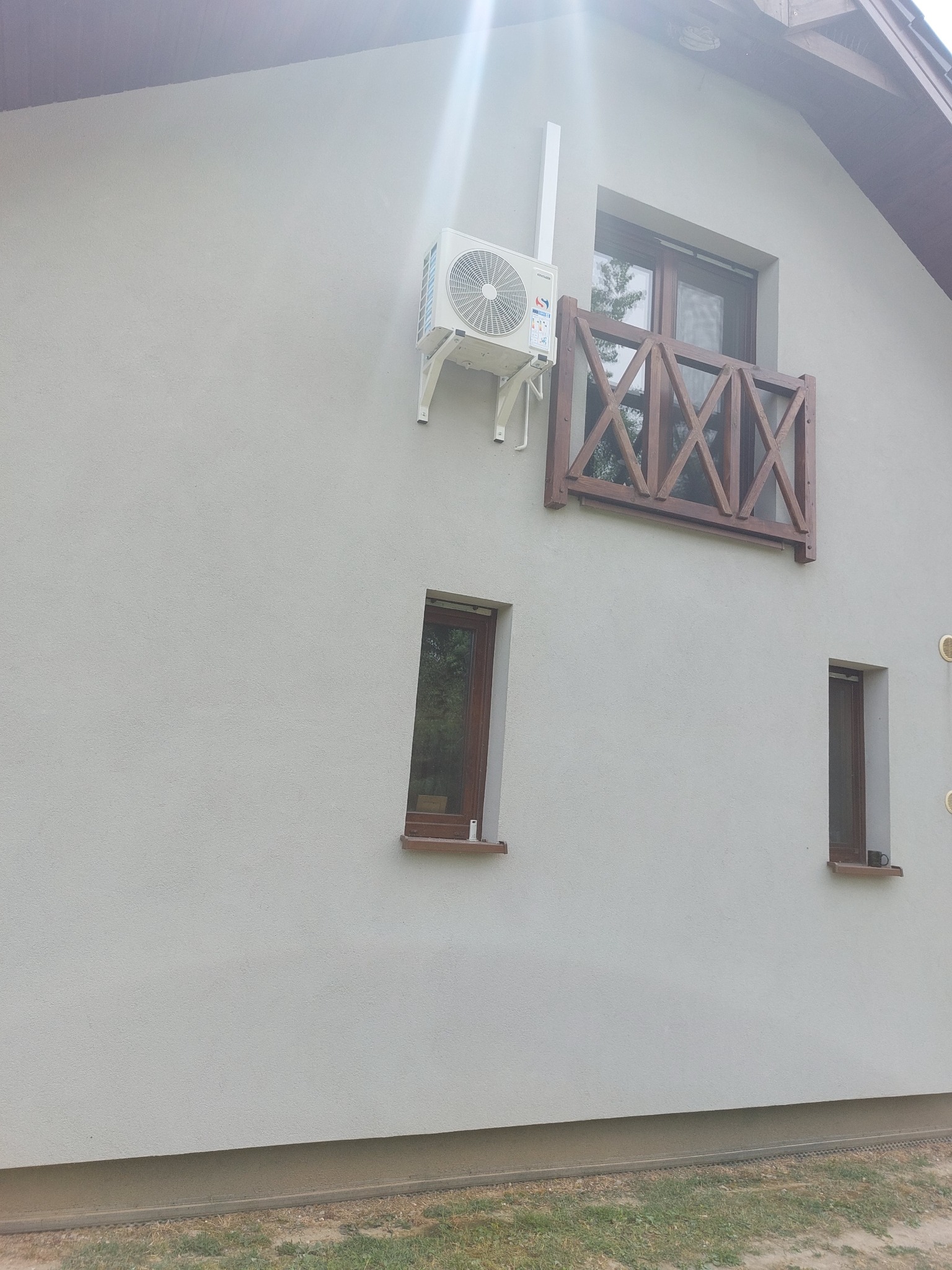 Biała jednostka zewnętrzna klimatyzacji zamontowana na ścianie domu z jasnym tynkiem, obok okna z drewnianą balustradą.