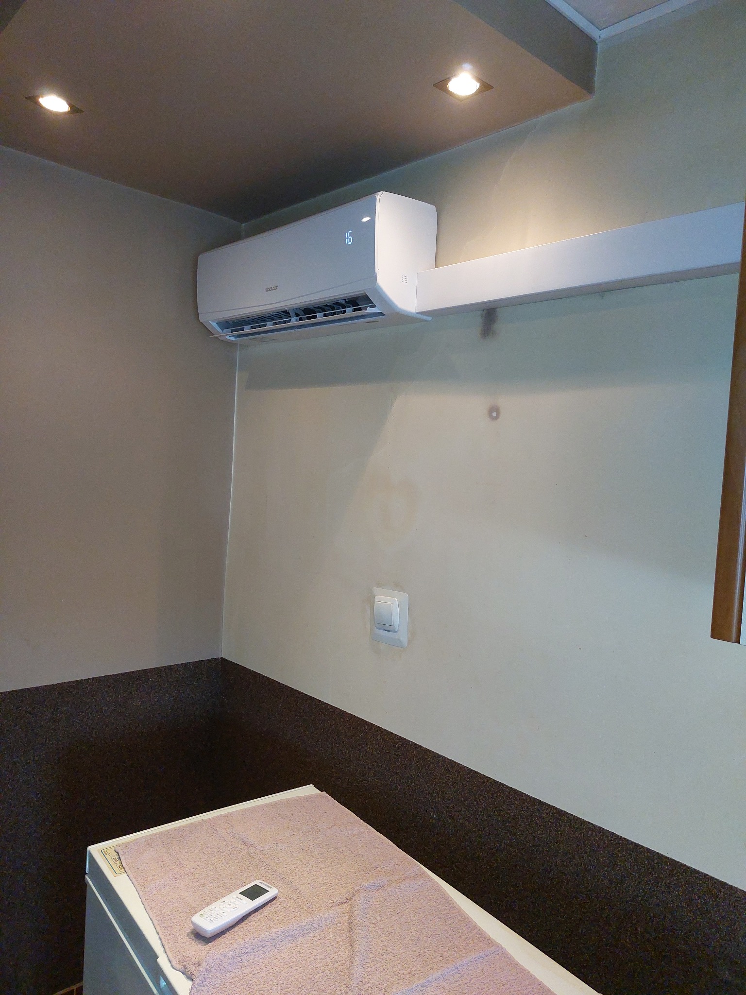 Biały klimatyzator z wyświetlaczem temperatury zamontowany na ścianie w pomieszczeniu o beżowych ścianach, widoczny pilot na stole przykrytym ręcznikiem.