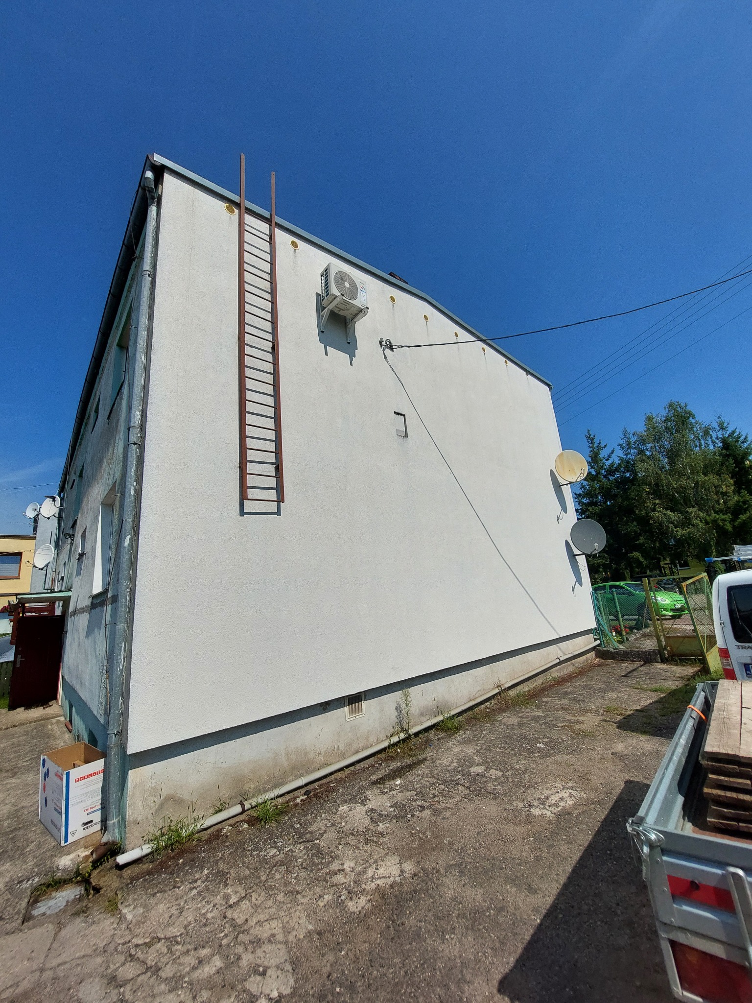Biały budynek z zamontowaną klimatyzacją, drabina przymocowana do ściany, trzy anteny satelitarne i przyczepka z deskami na zewnątrz, błękitne niebo w tle.