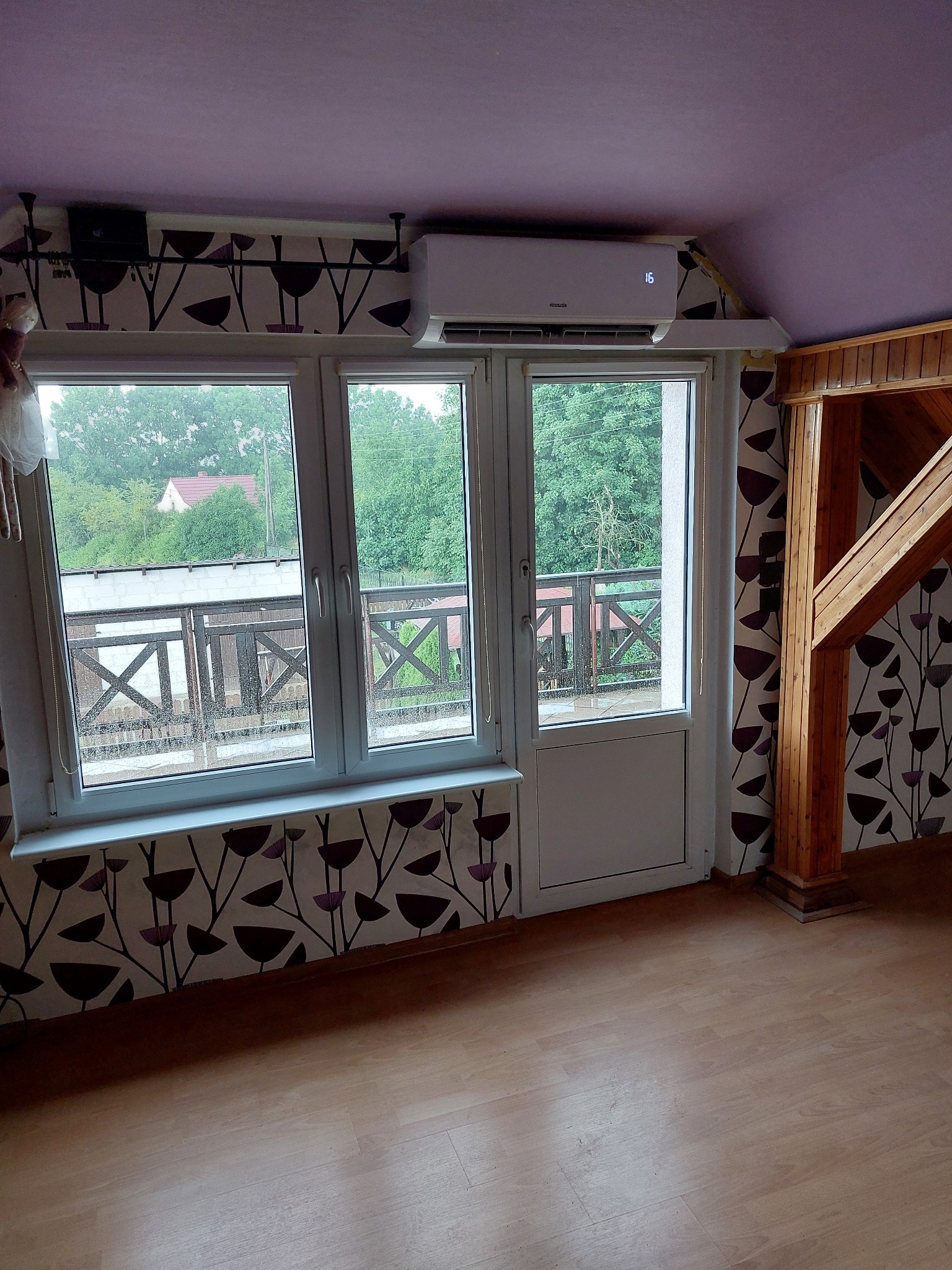 Biała jednostka klimatyzacyjna zamontowana nad oknem balkonowym w pokoju z wzorzystą tapetą i drewnianą konstrukcją. Widok z okna na zewnątrz.