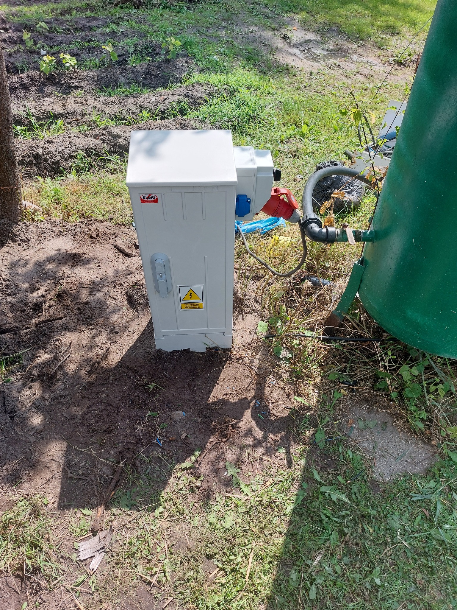 Zewnętrzna szafa elektryczna z ostrzeżeniem o wysokim napięciu, obok zielonego zbiornika, na trawiastym terenie z widocznymi przewodami i elementami instalacji.