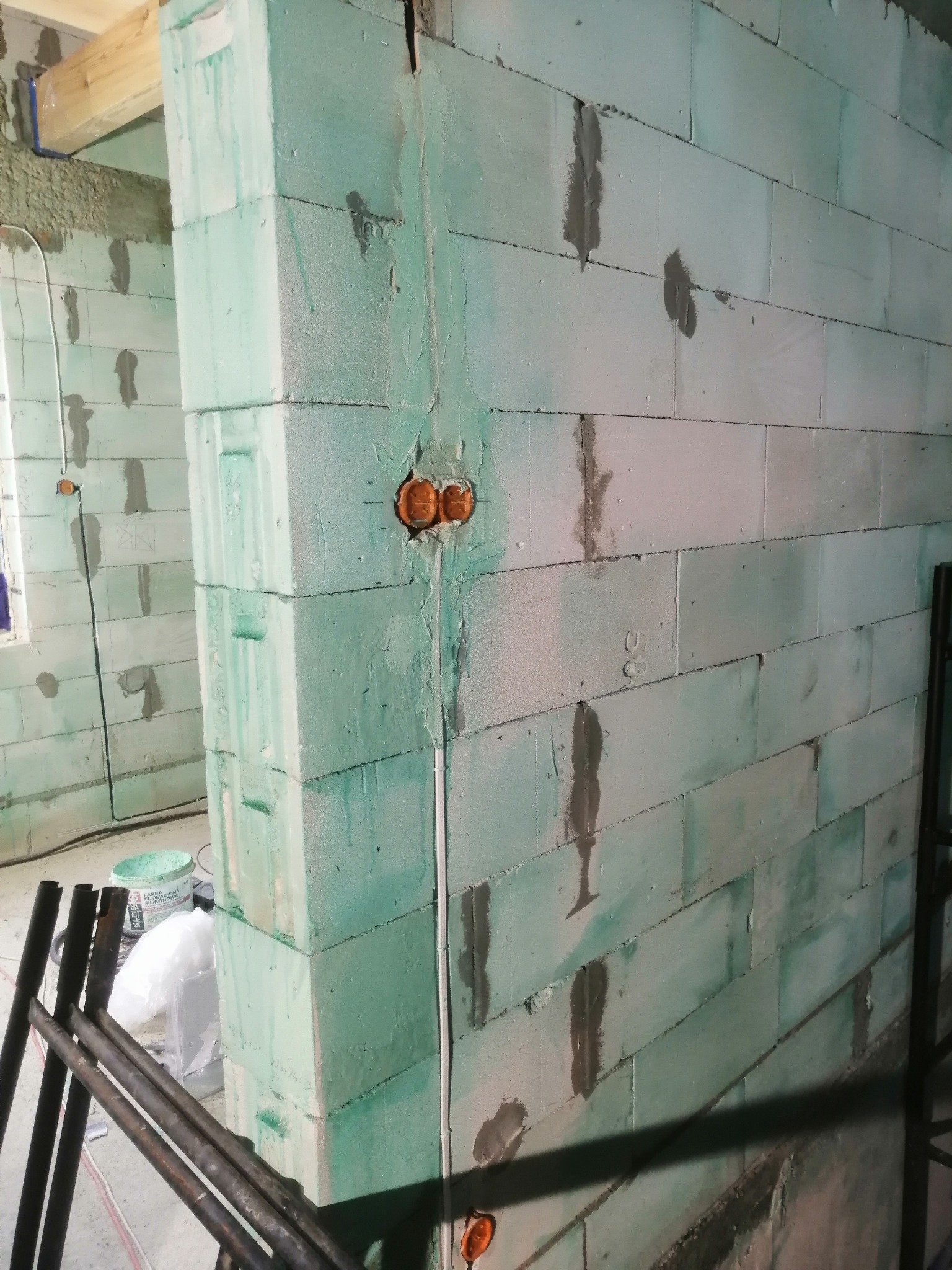 Surowy mur z betonu komórkowego w trakcie budowy, z widocznymi puszkami instalacyjnymi i przewodami elektrycznymi w bruzdach, częściowo pokryty zielonkawą farbą gruntującą.